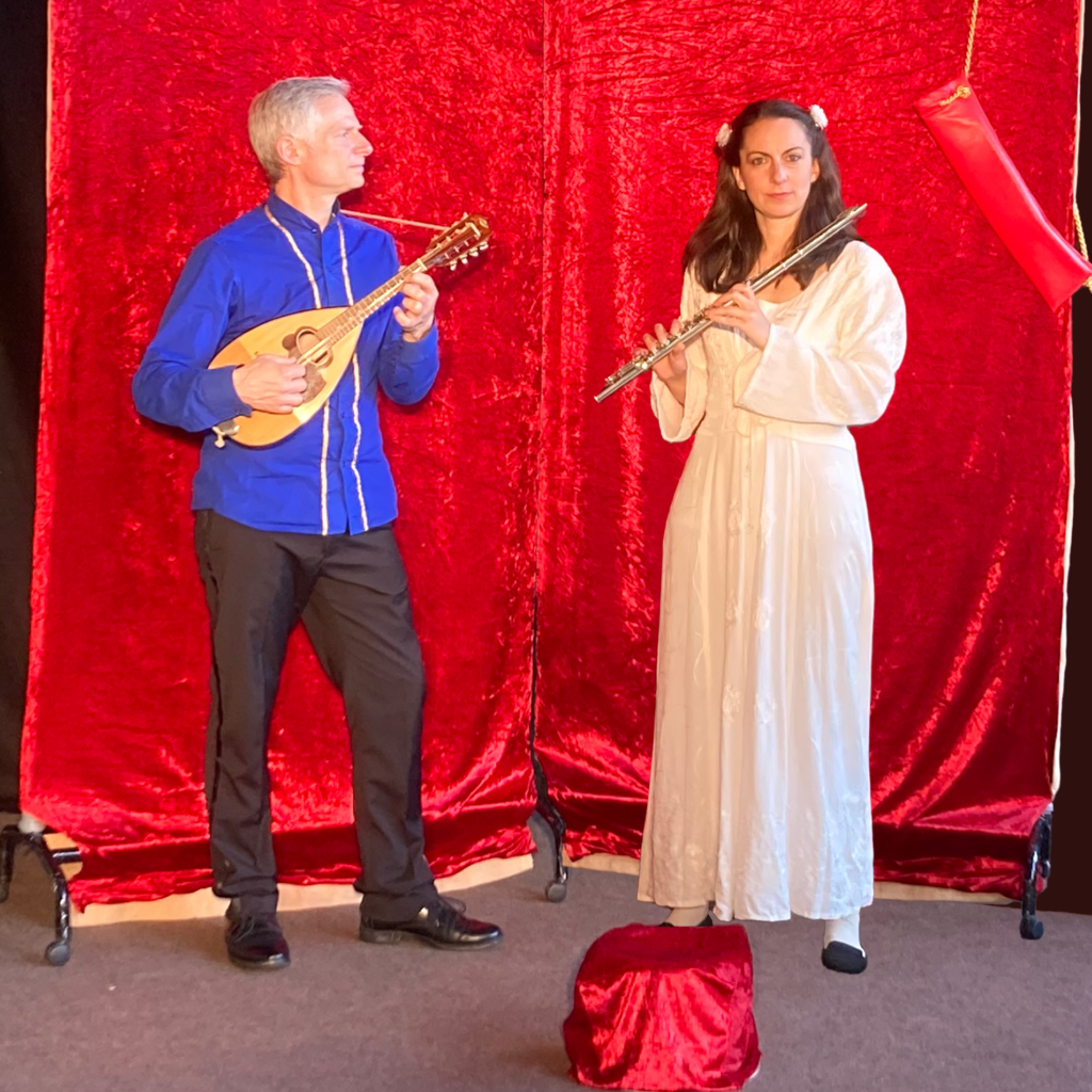 ein Mann mit blauer Kleidung und Mandoline schaut zu einer Frau im weißen Kleid, die Querflöte spielt